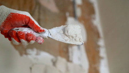Close-up of gloved hand holding putty knife with plaster, repairing damaged wall during home...