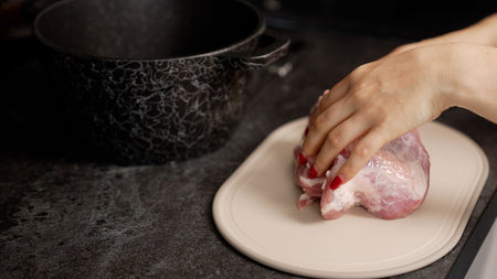 Professional female chef carefully cutting fresh pork meat with sharp knife on wooden cutting board in bright home kitchen, preparing ingredients for mealの写真素材