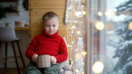 Cozy winter scene with child holding warm mug, gazing out window at snowy landscape, enjoying...