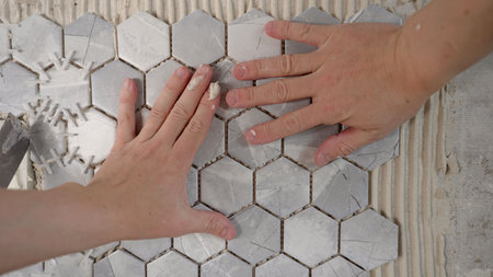 Construction workers are applying adhesive and pressing hexagonal tiles onto a wall, creating a...