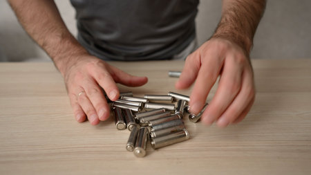 Close up of a man's hands checking a pile of used aa and aaa alkaline batteries on a wooden table for disposal and recyclingの写真素材