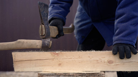 Skilled carpenter wearing gloves using an ax and a hammer to shape a wooden plank. Professional woodworking and construction processの写真素材