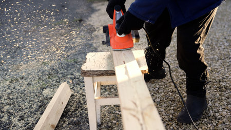 Close-up of a carpenter using an electric planer to smooth a wooden plank. Sawdust flies as the...