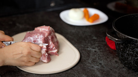 Hands preparing raw pork meat on a cutting board, ready for cooking with vegetables in a kitchen...