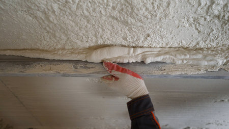 A construction worker in gloves applies a thick white plaster mix along a metal guide on a wall,...