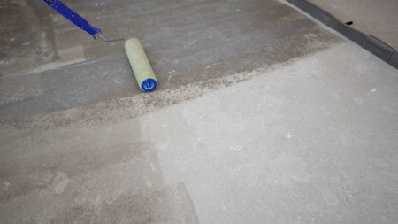 A close-up shot shows a paint roller applying a light grey primer or paint to a concrete floor, demonstrating the process of floor preparation and renovation. The roller moves steadily,の写真素材