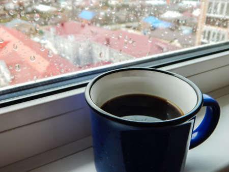 a blue mug with coffee stands by the window, and outside it is rainingの写真素材