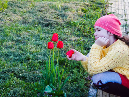 A little girl in a yellow sweater and a red hat sits on the grass and holds a bouquet of tulips.の写真素材