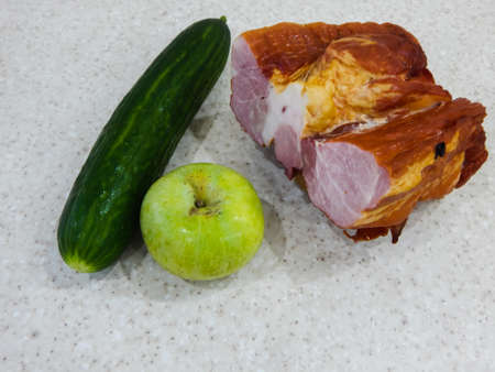 a piece of smoked pork, a green cucumber, an apple lie side by side on a white backgroundの写真素材