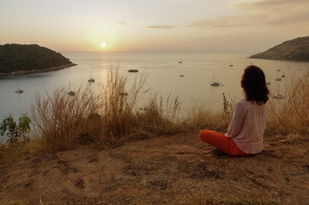 Young girl sitting in yoga lotus meditation position front to seaside on the rocks an watching the sun goes down in a golden hour. sunset over sea.の写真素材