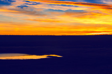 Beautiful sunset over a field with a lake in the foreground. The sky is filled with orange and pink...