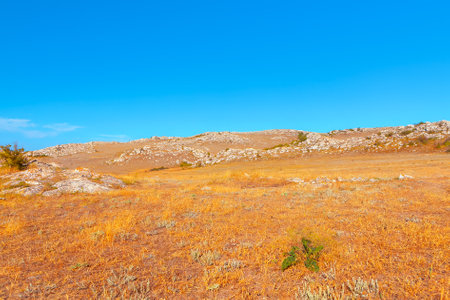Barren rocky hillside with a hill on background. Blue sky over the hill with dry grassの写真素材