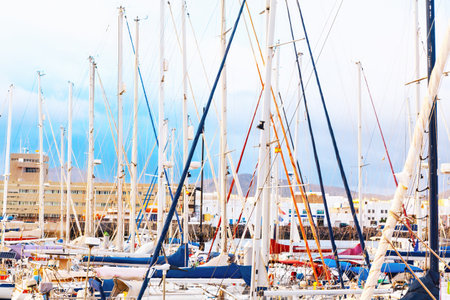 Densely packed marina with numerous sailboats docked closely together. Masts of the sailboats create a complex and visually striking pattern against the backdrop of buildings and a cloudy skyの写真素材