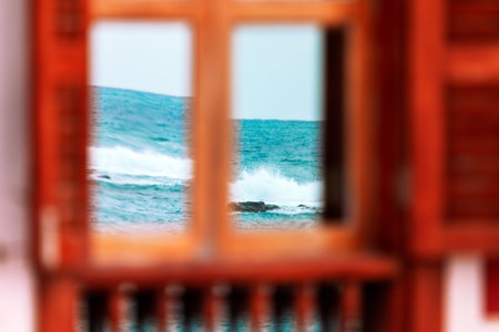 Ocean view framed through a wooden window, with its vibrant waves crashing in the distance. Foreground features blurred elements of the wooden window, contrasting with vivid of blue seaの写真素材