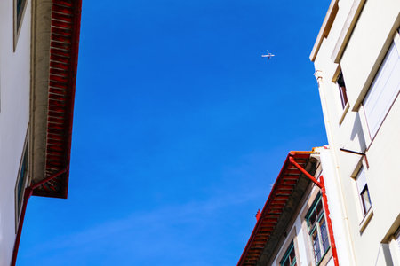 Clear blue sky is framed by roofs of two buildings on either side. Airplane is visible in distance flying high above. Buildings have red roofs and white walls, creating contrast with vibrant blue skyの写真素材