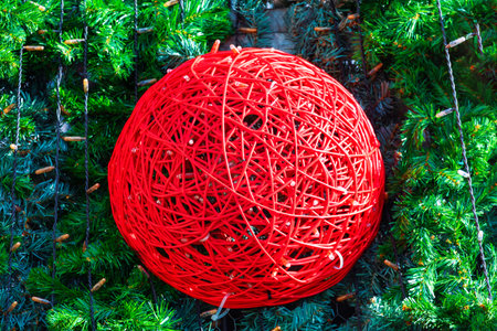Spherical decoration made of red intertwined wires, set against a backdrop of green pine branches....