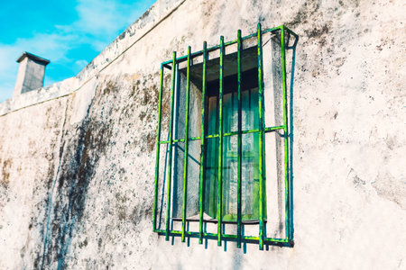 Rectangular window with greenish glass panes is set in weathered, white wall. Window is protected by metal grille with vertical and horizontal bars, which are slightly rusted and have a greenish tintの写真素材