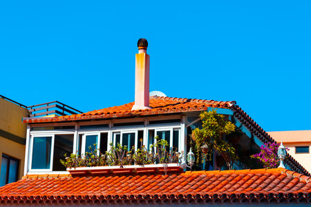 Upper part of a house with a tiled roof, featuring a chimney and a balcony adorned with plants and flowers. Blue sky in background contrasts with the warm colors of the roof and greeneryの写真素材