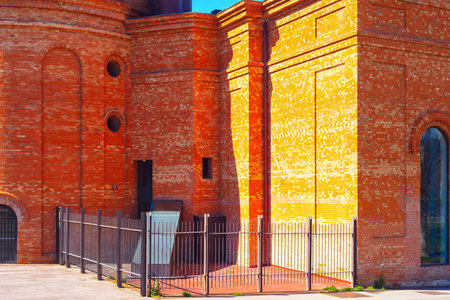 Red brick building with intricate architectural details, arched windows and fence in foreground. Structure is illuminated by sunlight highlighting its texture and warm tones. Urban and historical themeの写真素材
