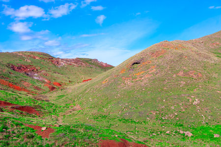 Scenic landscape with rolling hills covered in green grass and patches of red soil under a bright blue sky. Green vegetation and the red soil, along with the clear sky makes scene visually strikingの写真素材
