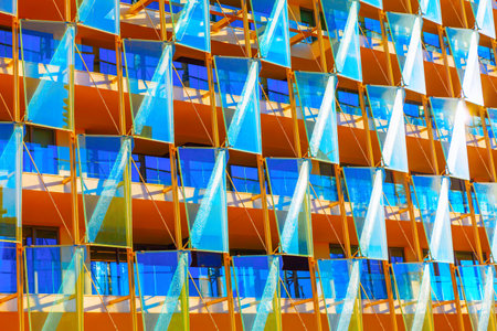 Modern building facade with pattern of glass panels arranged in a geometric design. Panels are angled, allowing reflections of sky and a play of light, while balconies behind are visible through glassの写真素材
