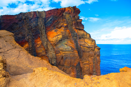 Striking cliffside with vibrant, multicolored rock layers overlooking a deep blue ocean under a partly cloudy sky. Rugged textured cliff and smooth expanse of sea makes scene visually captivatingの写真素材