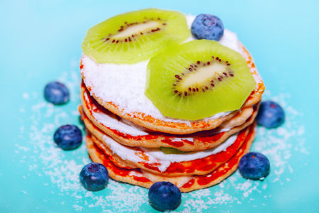 Stack of pancakes topped with powdered sugar and surrounded by blueberries on a light blue surface.の写真素材