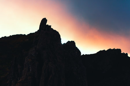 Silhouette of a black rocky mountain against a cloudy sky, with the sun partially obscured behind...