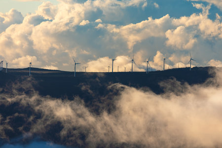 Wind turbines stand tall on a misty mountain ridge, partially veiled by low-hanging clouds. Dramatic...