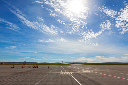 Wide view of an empty airport runway under a bright sky. There are a few pieces of ground equipment and traffic cones visible on tarmac, with vast expanse of open land and distant hills in backgroundの写真素材