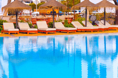 Poolside area with beige lounge chairs arranged in a row under thatched umbrellas, reflecting in...
