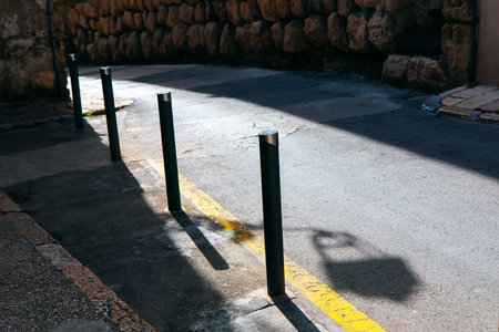 Narrow street illuminated by sunlight, highlighting bollards lined up along road marking. On one side, textured stone wall contrasts with smooth pavement, while shadows add depth to urban compositionの写真素材