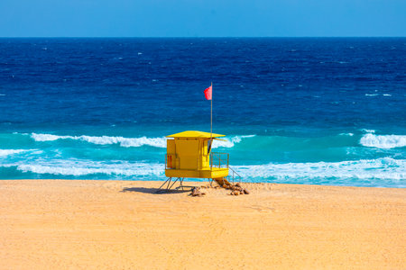 Bright yellow lifeguard tower stands alone on a sunlit sandy beach, with turquoise waves crashing under a vivid blue sky. A red flag flutters, signaling caution amid the serene coastal beautyの写真素材