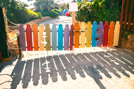 Small colorful wooden gate made of vertical planks painted in different bright colors, some featuring anchor cutouts. Sunlight casts long, distinct shadows of the gate on the cobblestone path belowの写真素材