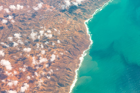 Aerial view captures a rugged, mountainous coastline meeting deep blue ocean under a partly cloudy sky. Contrast between arid, rocky terrain and vibrant sea highlights dramatic beauty of landscapeの写真素材