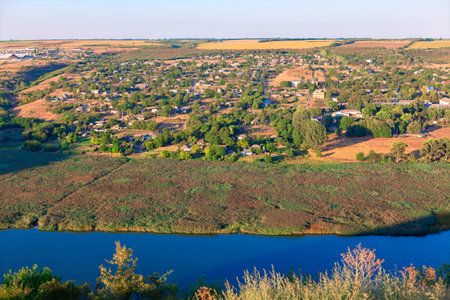 Village in Moldova along Dniester riverside, with scattered houses and patches of greenery spread across gently rolling landscape. Calm blue waters of Dniester River flow bordered by dense vegetationの写真素材
