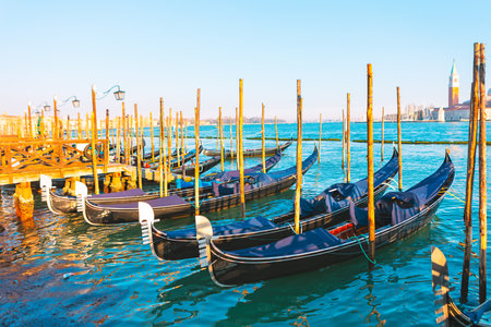 Traditional gondolas moored along a wooden pier in Venice, Italy, bask under the warm morning sun. Calm turquoise waters and a distant bell tower complete this iconic Venetian sceneの写真素材