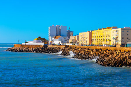 Vibrant coastal scene in Cadiz, Andalusia Spain, with waves crashing against a sturdy stone breakwater along shoreline. Colorful buildings and modern high-rise line background under blue skyの写真素材