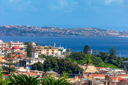 Picturesque view of Messina, Sicily, with colorful buildings along coastline and vibrant sea stretching toward horizon. In background, rolling hills dotted with homes enhance charming coastal townの写真素材