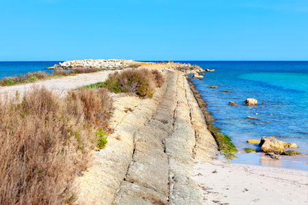 Coastal pathway meanders along a stone seawall, leading to the tranquil turquoise waters. Pathway lined with a stone seawall, leading to a tranquil beach and the deep blue seaの写真素材