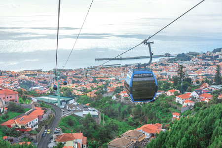 In Funchal cable car glides over a vibrant town with red-roofed houses, set against lush green hillside and the sparkling ocean. Scenic view captures charm of this coastal city on Madeira Islandの写真素材
