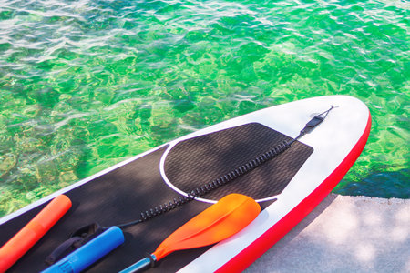 Paddleboard rests beside crystal clear water, holding paddles and a coiled leash. Water sports...
