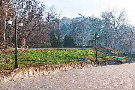 Quiet park scene features a paved walkway with vintage style lamp posts and a green bench....