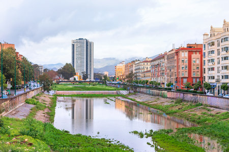 View of Malaga shows a river with reflections, surrounded by colorful historic buildings and a modern tower in distance. Cloudy sky and mountains create a striking urban and natural contrastの写真素材