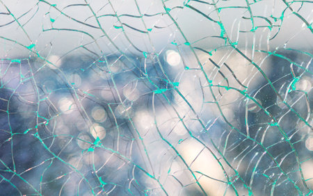 Sheet of shattered glass, surface covered in a network of sharp cracks. Blurred background reveals...