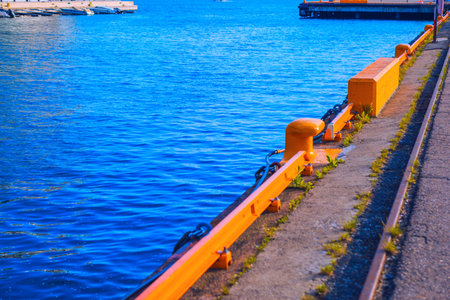 Calm blue body of water next to a concrete pier with a bright orange railing and mooring bollard. The pier has railroad tracks along its edge, with some overgrown weedsの写真素材