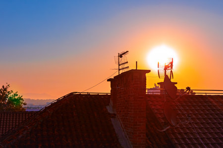 Vibrant sunset casts orange glow over tiled rooftop, silhouetting a brick chimney and TV antenna against deepening sky. Distant hills and sparse trees frame scene with  sun raysの写真素材