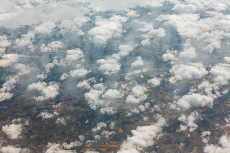 Aerial photo from an airplane window shows a patchwork of farmland fields stretching to horizon. Scattered fluffy white cumulus clouds partially obscure terrain below vast partly cloudy skyの写真素材