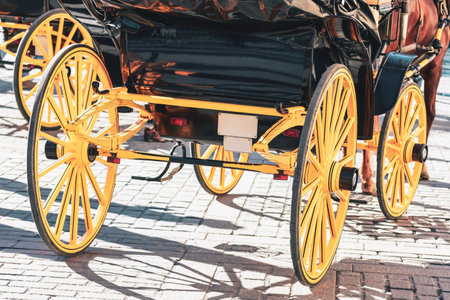 View of the rear wheels and undercarriage of a traditional black horse-drawn carriage, featuring bright yellow wooden spokes. Wheels of horse-drawn transport onto textured brick pathの写真素材
