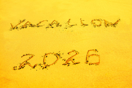 Word VACATION is inscribed into sand with year "2026" written below. VACATION 2026 etched into smooth beach sand, hinting future travel plans. Golden sand texture evoke leisure and coastal atmosphereの写真素材